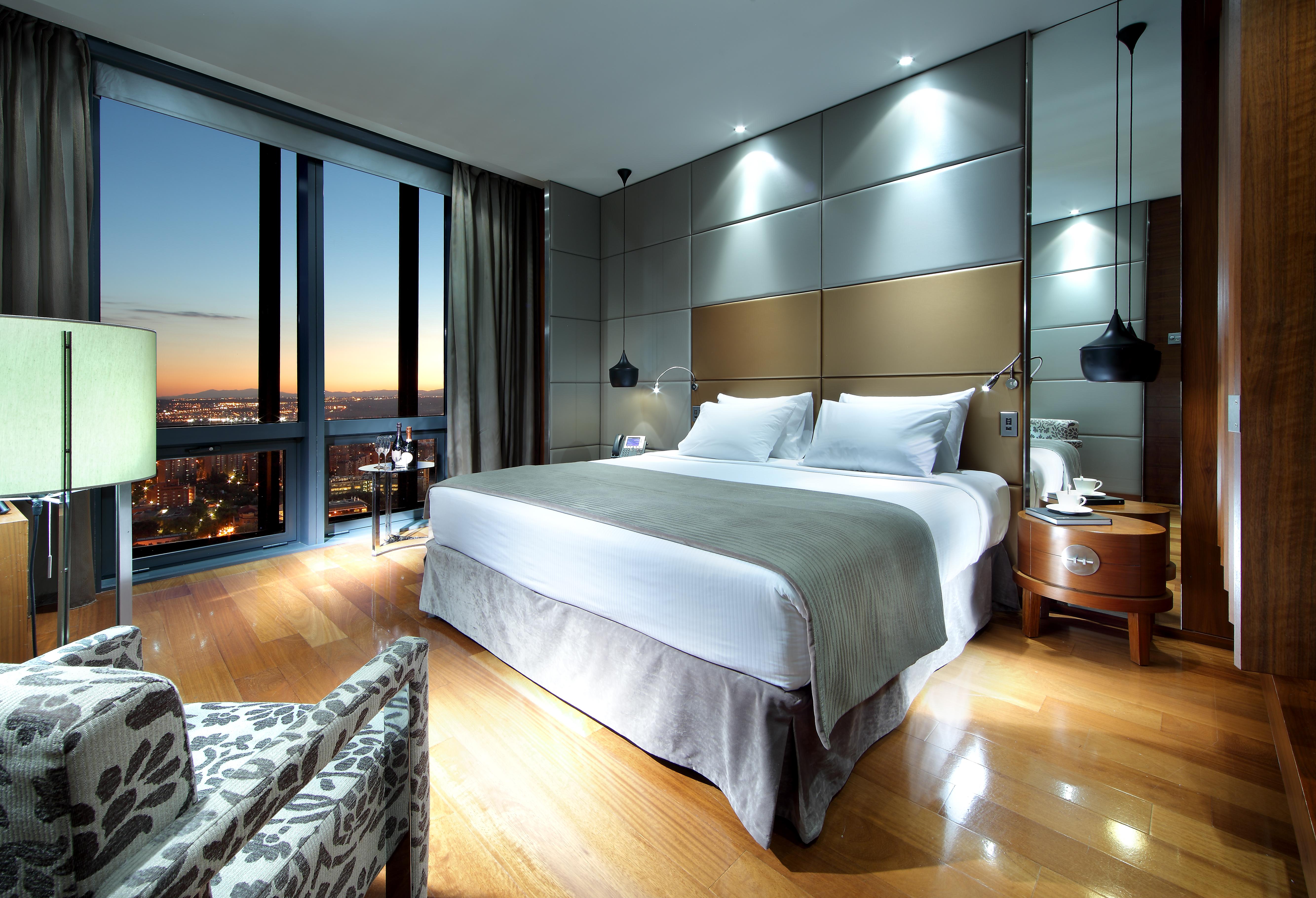 Eurostars Madrid Tower Hotell Exteriör bild The image shows a modern hotel room featuring a large bed with white linens and a gray throw. There are stylish bedside lamps and a sleek nightstand. A comfortable chair with a patterned fabric sits to the side. The room has large windows that reveal
