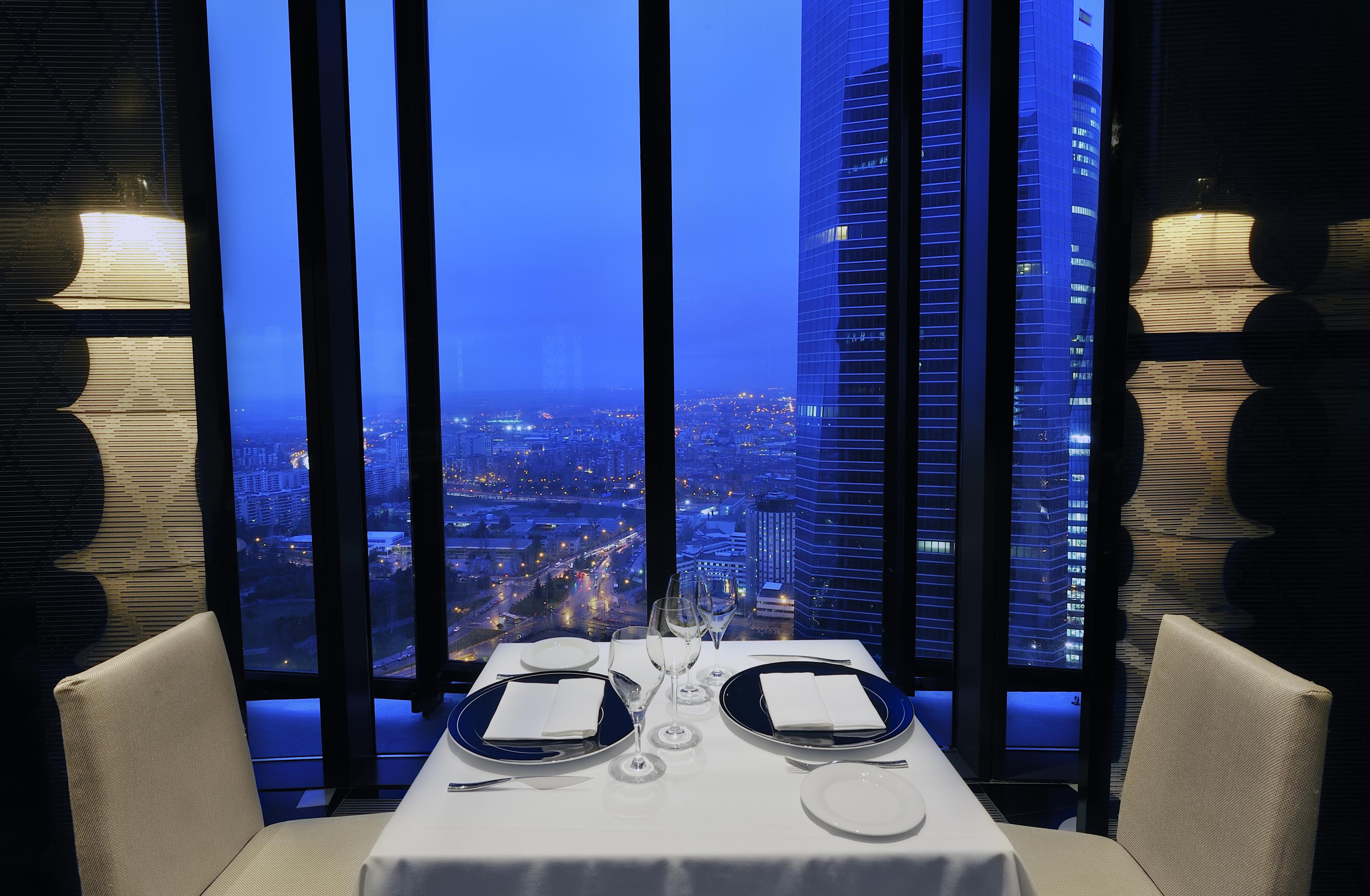 Eurostars Madrid Tower Hotell Restaurang bild The photo shows a fine dining restaurant setting with a table for two, elegantly set with white tablecloth, plates, glasses, and utensils. The table is positioned near large windows that offer a panoramic view of a cityscape at dusk, illuminated by l