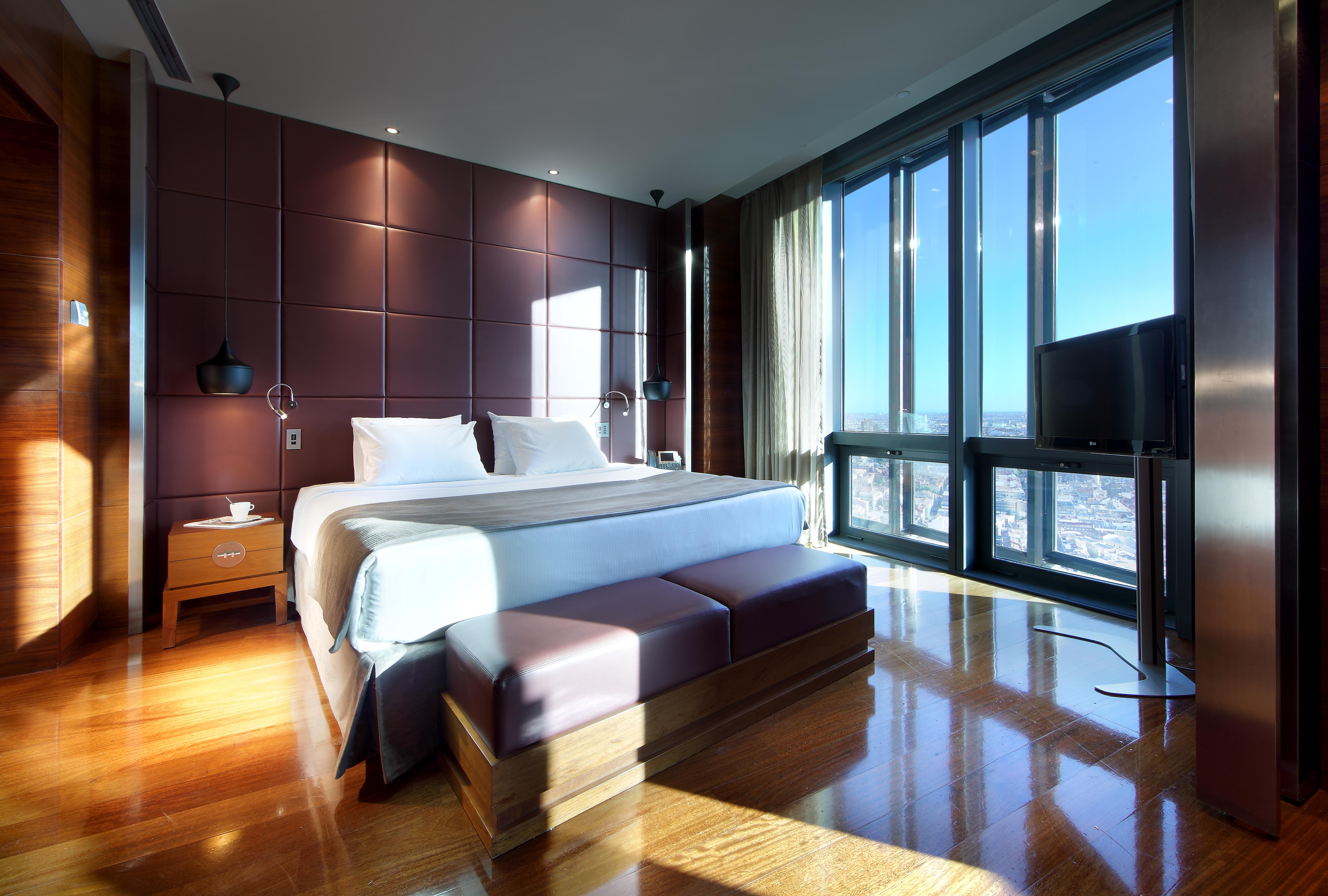 Eurostars Madrid Tower Hotell Exteriör bild The photo shows a modern hotel room with sleek design features. It includes a large bed with white bedding, positioned against a wall with dark, textured panels. On either side of the bed, there are bedside tables with lamps. A stylish bench sits at 