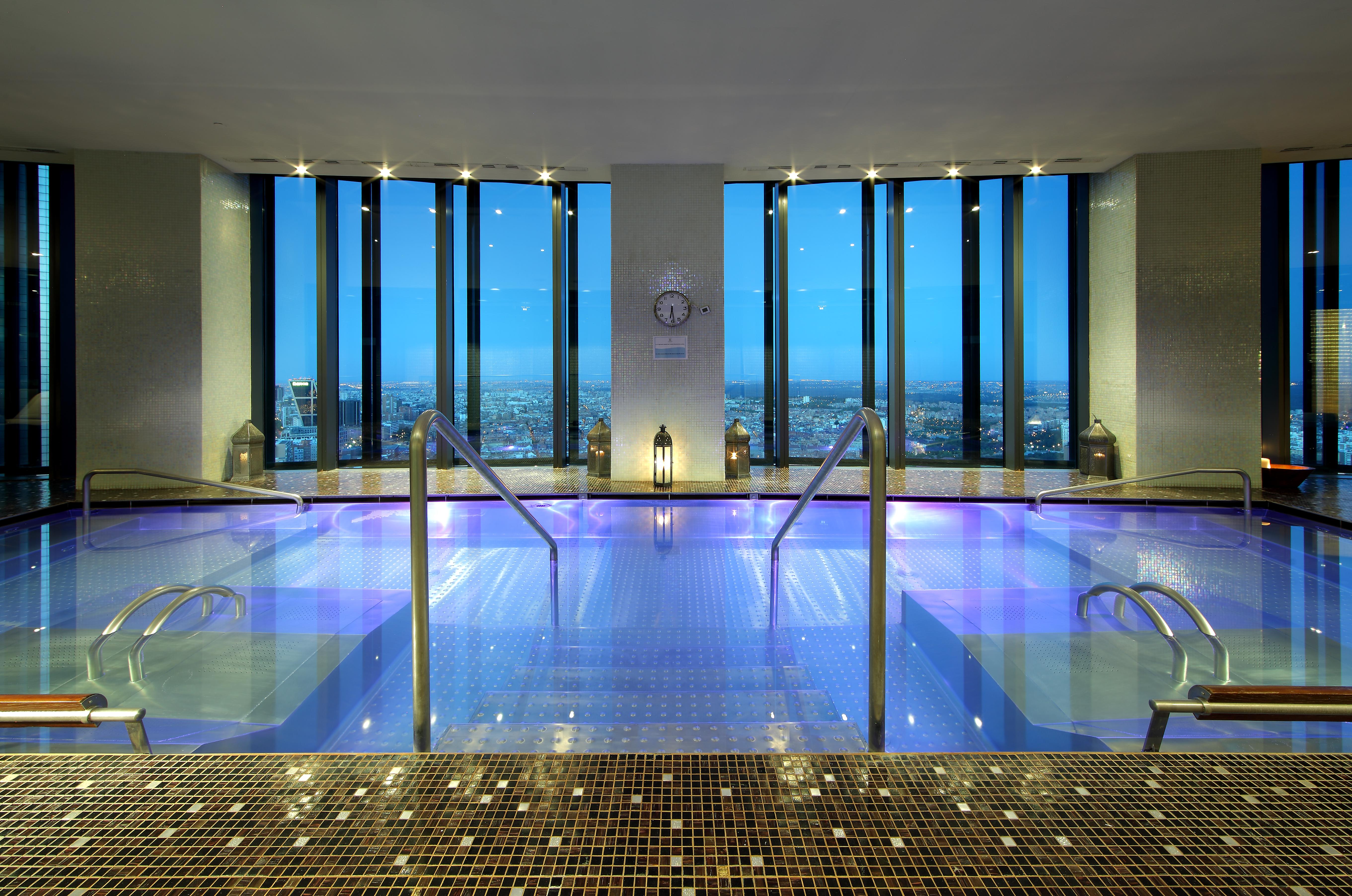 Eurostars Madrid Tower Hotell Exteriör bild The image shows a luxurious spa area with a large, tranquil pool that has a vibrant blue hue. The pool is surrounded by sleek, modern design elements, including steps leading down into the water. Large windows provide a stunning view of the cityscape