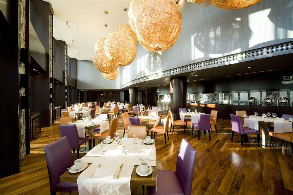 Eurostars Madrid Tower Hotell Restaurang bild The photo shows a modern restaurant interior featuring elegant decor. The dining area includes tables set for guests with white tablecloths, plates, and cutlery. The seating consists of a mix of wooden chairs, some upholstered in purple fabric. The l