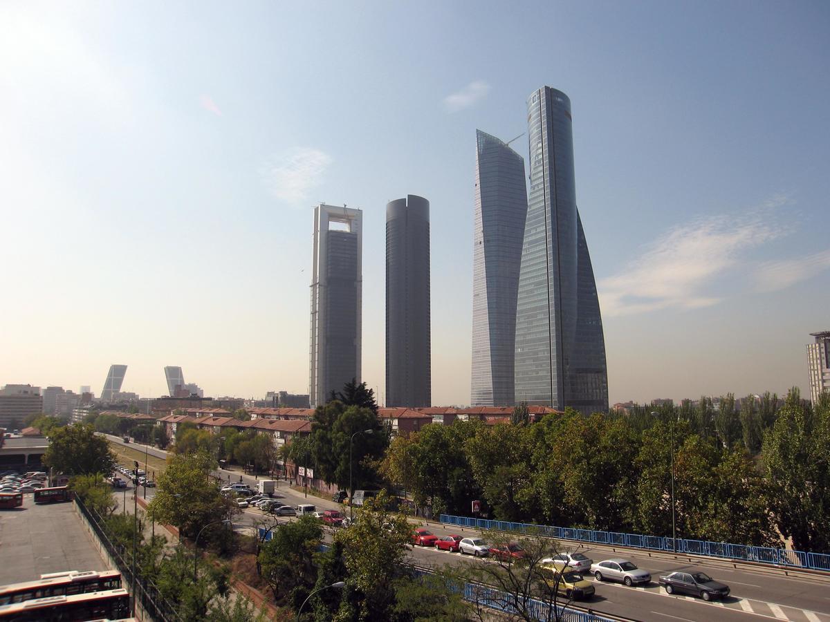 Eurostars Madrid Tower Hotell Exteriör bild The photo shows a cityscape featuring several modern skyscrapers, prominently four tall buildings in the foreground. The structures have distinctive architectural designs, with varying shapes and heights. The scene includes a tree-lined area and a bu