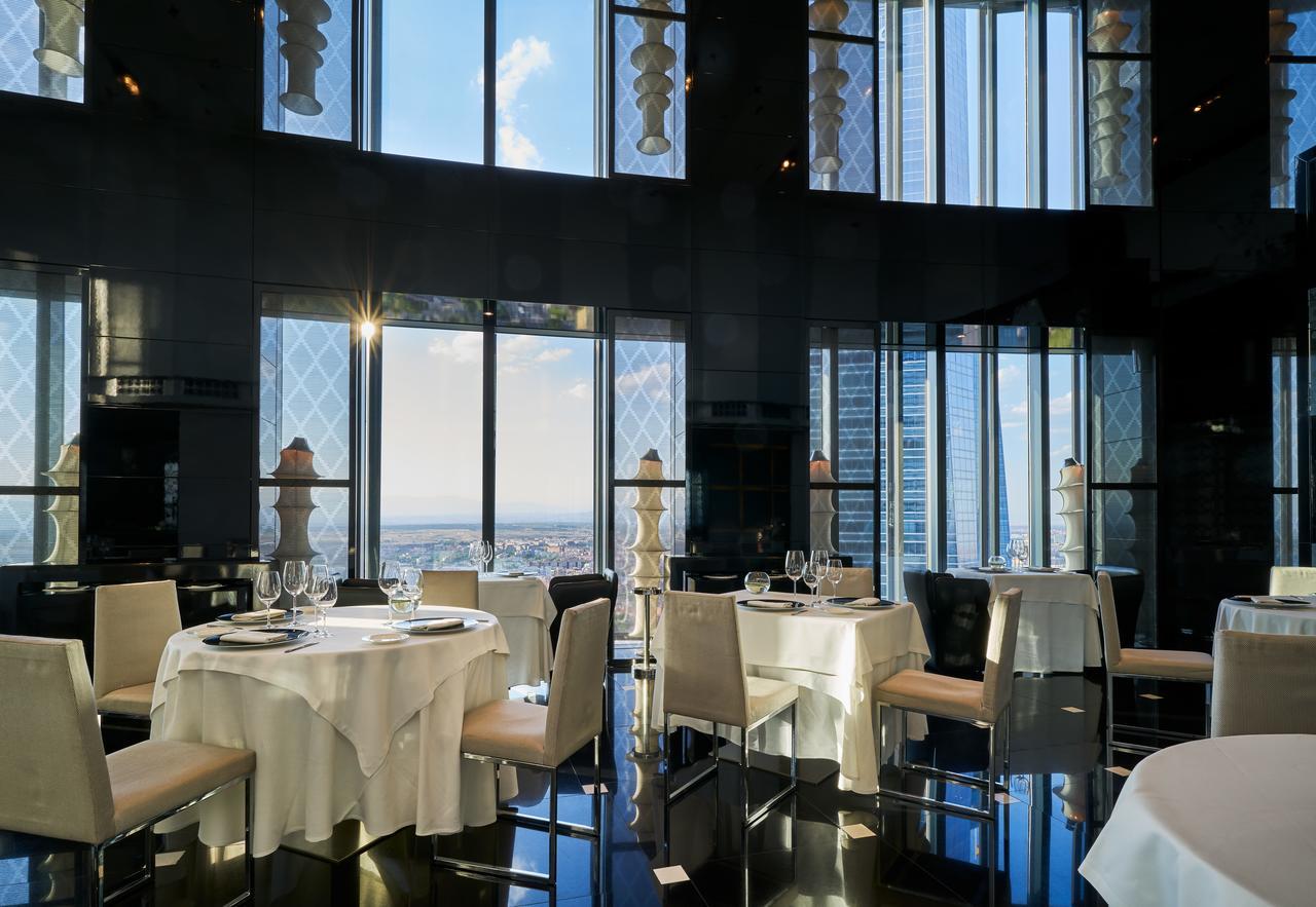 Eurostars Madrid Tower Hotell Exteriör bild The photo features an elegant restaurant interior with large windows that provide a panoramic view of the city skyline. The tables are elegantly set with white tablecloths and fine dining tableware, including glasses and plates. The decor appears mod